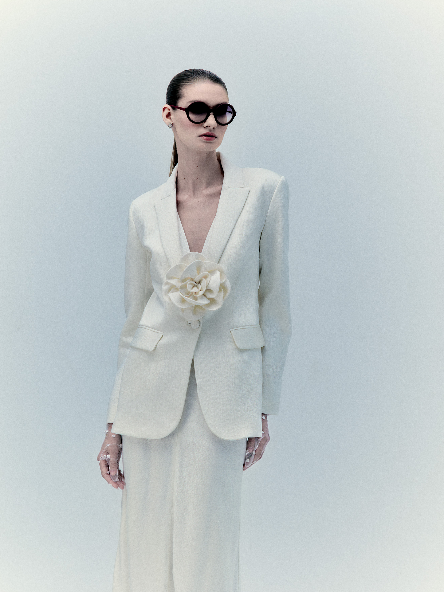 A person wearing the Daniella Dress in white, featuring a large flower detail, and sporting sunglasses, stands against a plain background to showcase this exclusive piece. Inquire for more details or further availability upon request.