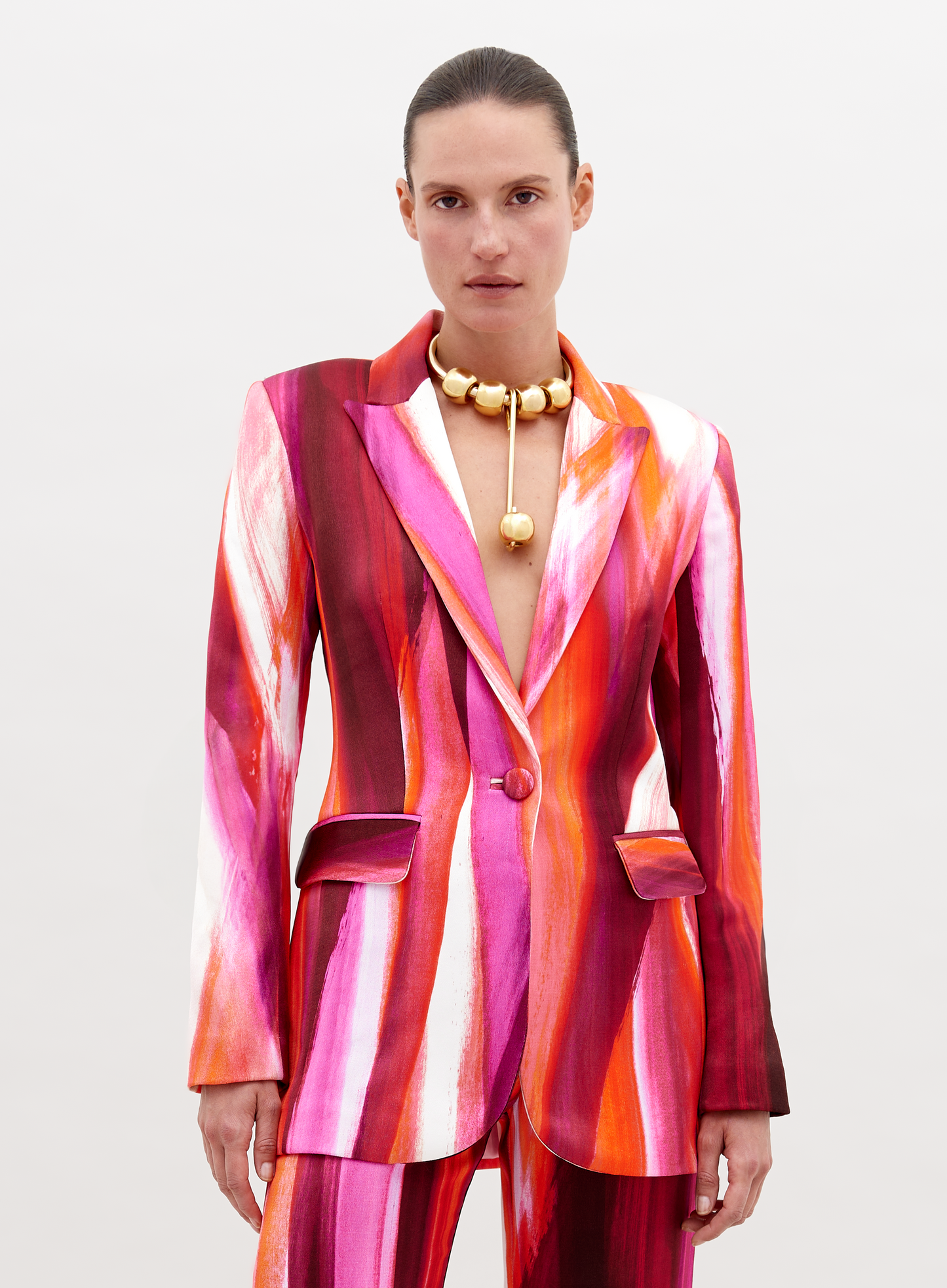 Woman wearing a vibrant, multi-colored striped Rebeca Jacket Magenta Abstract Waves with a deep neckline, accessorized with a chunky gold choker necklace, standing against a plain white background. Pre-order now and have it ship by date of November 15th, 2024.