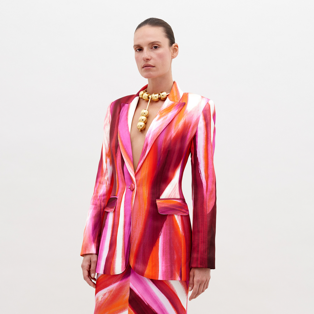 Woman wearing a vibrant, multi-colored striped Rebeca Jacket Magenta Abstract Waves with a deep neckline, accessorized with a chunky gold choker necklace, standing against a plain white background. Pre-order now and have it ship by date of November 15th, 2024.