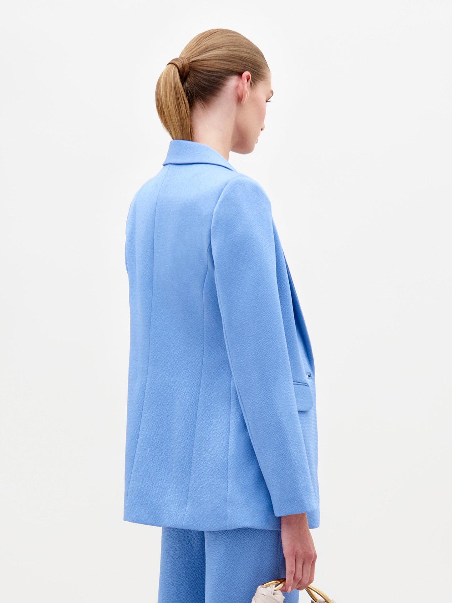 A person wearing the Rebeca Jacket in Periwinkle over a knotted shirt and trousers stands against a plain white background, holding glasses in their right hand. Pre-order now to ensure it arrives by February 15th, 2025!