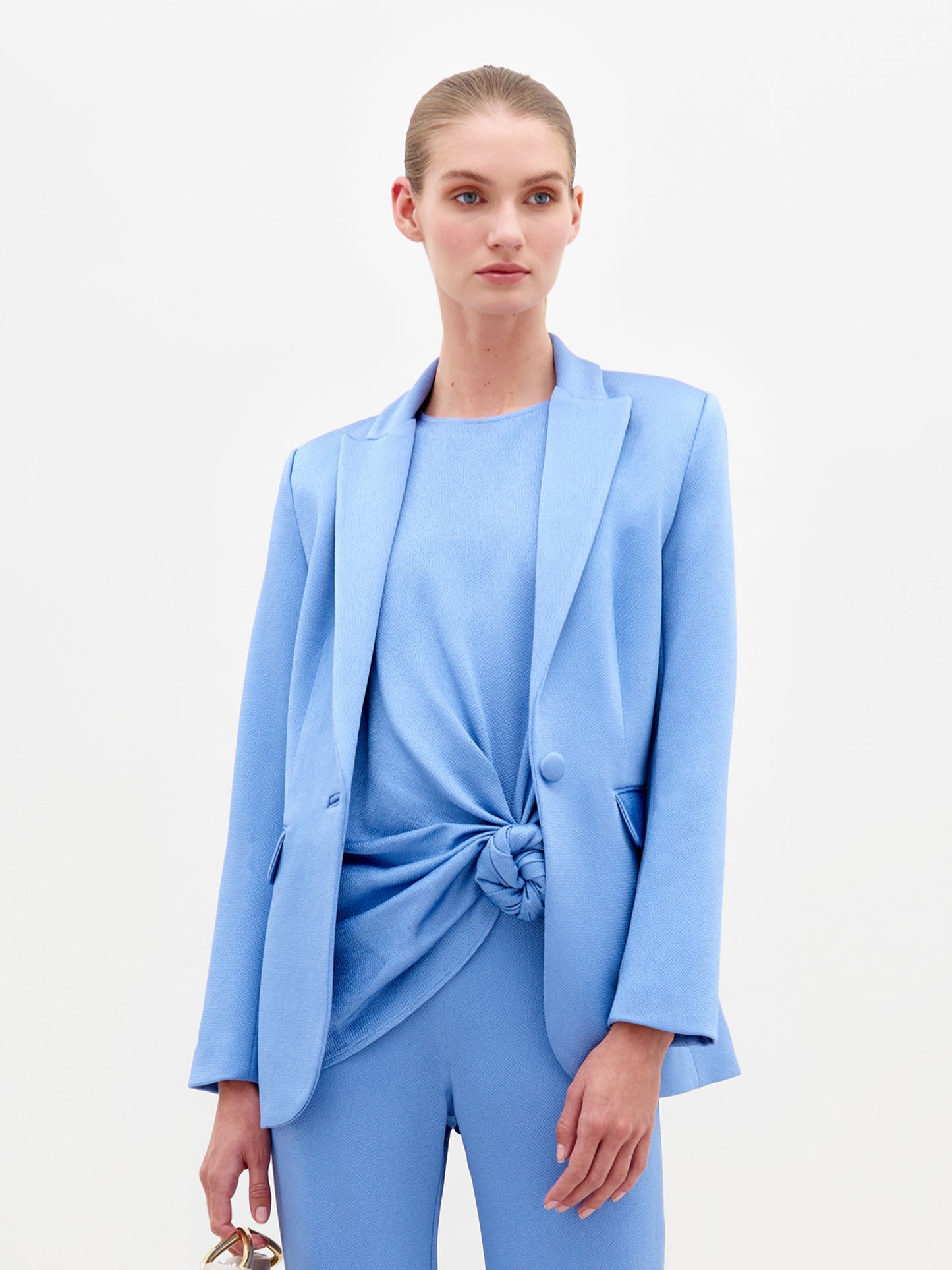 A person in a blue Rebeca Jacket with a single-button closure and sandals, holding a beige bag, stands against a plain white background, exuding the elegance of fine Italian textile.