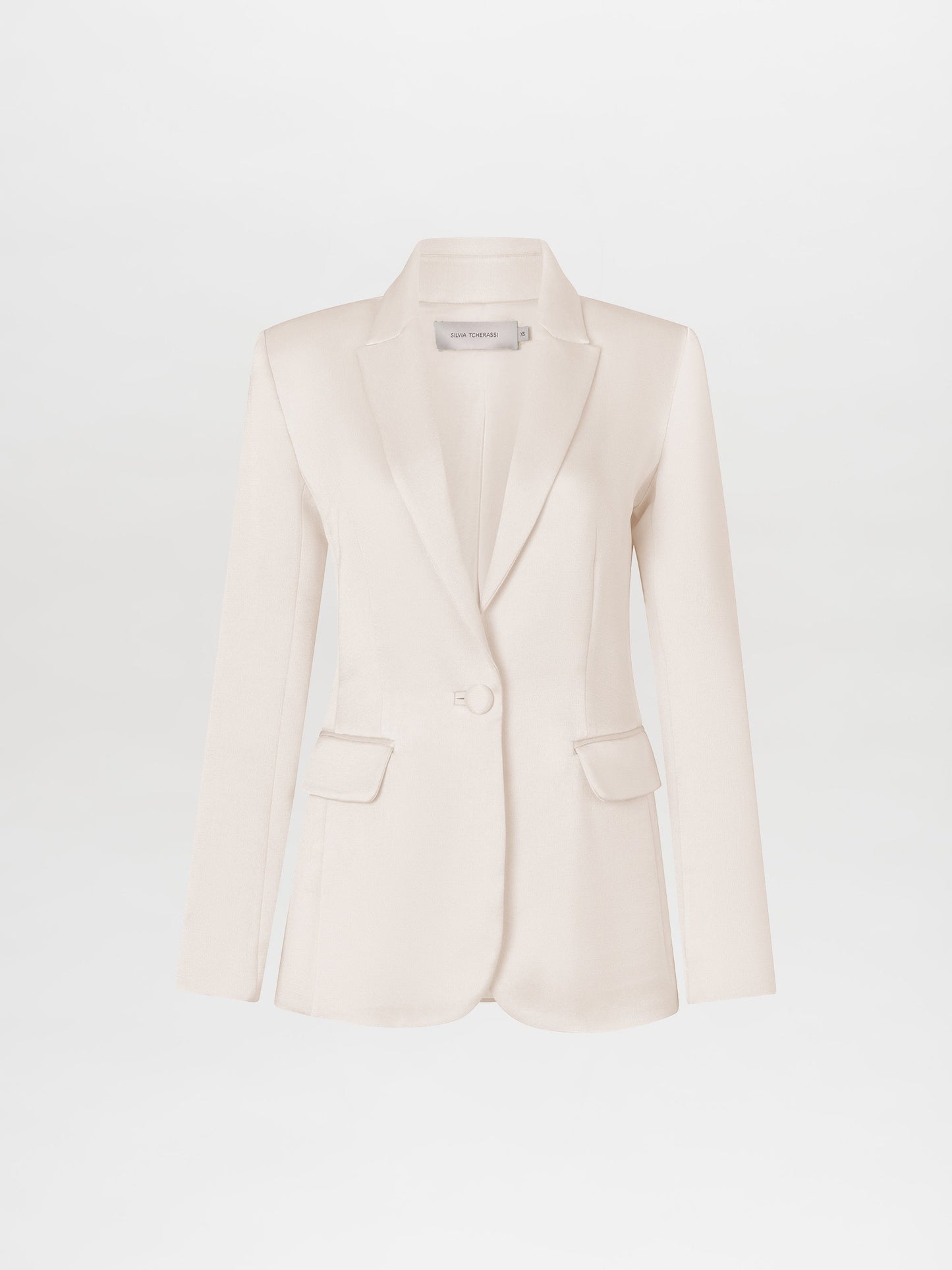 A person with a neutral expression is wearing the Rebeca Jacket White and white pants, standing against a plain background with arms crossed, available for pre-order starting February 15th, 2025.