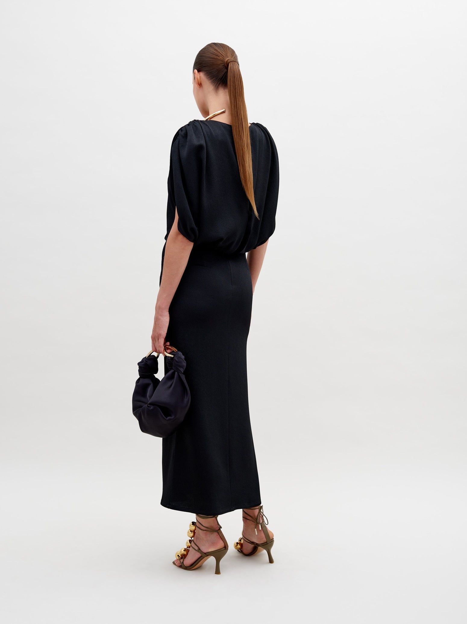 A person in the Reese Dress, a stunning black hammered satin piece with a decorative gold neckline, holds a chic black round handbag and wears gold-embellished high heels, standing elegantly against a plain white background.