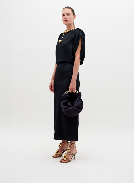 Against a plain background, a person stands elegantly in the Reese Dress Black with gold accessories, holding a matching black handbag. They also sport high-heeled sandals adorned with gold embellishments. Pre-order now to ensure your look is ready by the ship-by date of November 15th, 2024.