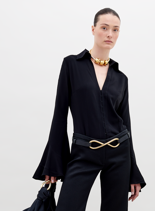 A person with slicked-back hair wears a black outfit with flared sleeves, a gold necklace, and a belt featuring a large gold infinity symbol. 