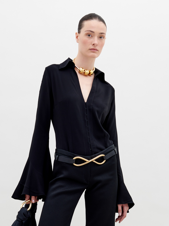 A person with slicked-back hair wears a black outfit with flared sleeves, a gold necklace, and a belt featuring a large gold infinity symbol. They hold the Reggie Blouse Black in their hand, available for pre-order now and set to ship by date November 15th, 2024.