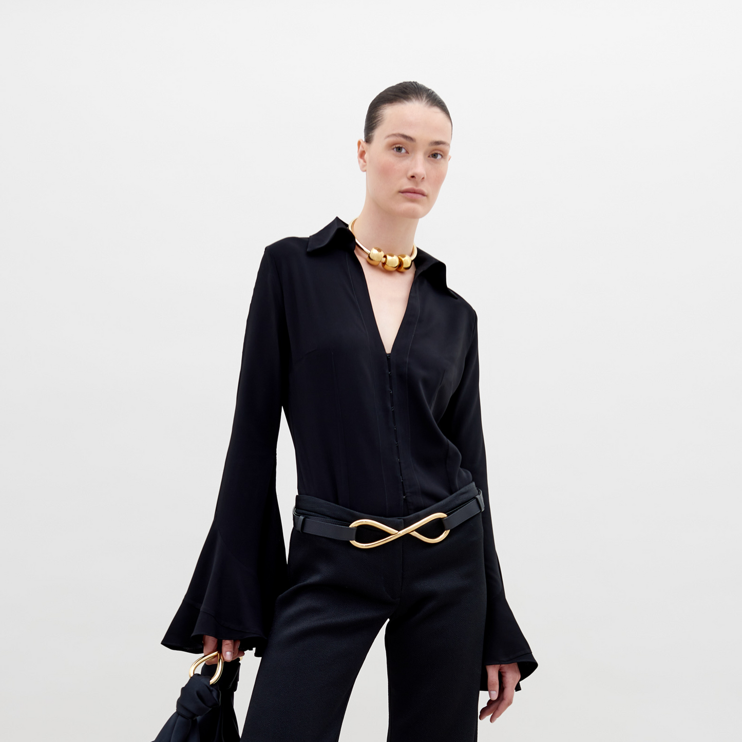 A person with slicked-back hair wears a black outfit with flared sleeves, a gold necklace, and a belt featuring a large gold infinity symbol. They hold the Reggie Blouse Black in their hand, available for pre-order now and set to ship by date November 15th, 2024.