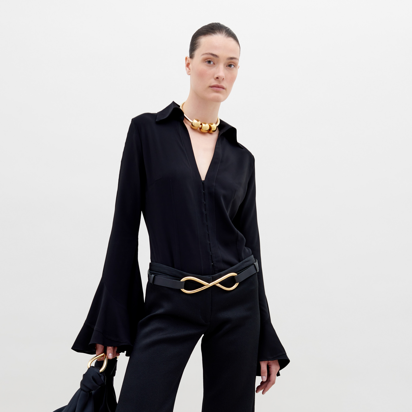 A person with slicked-back hair wears a black outfit with flared sleeves, a gold necklace, and a belt featuring a large gold infinity symbol. They hold the Reggie Blouse Black in their hand, available for pre-order now and set to ship by date November 15th, 2024.