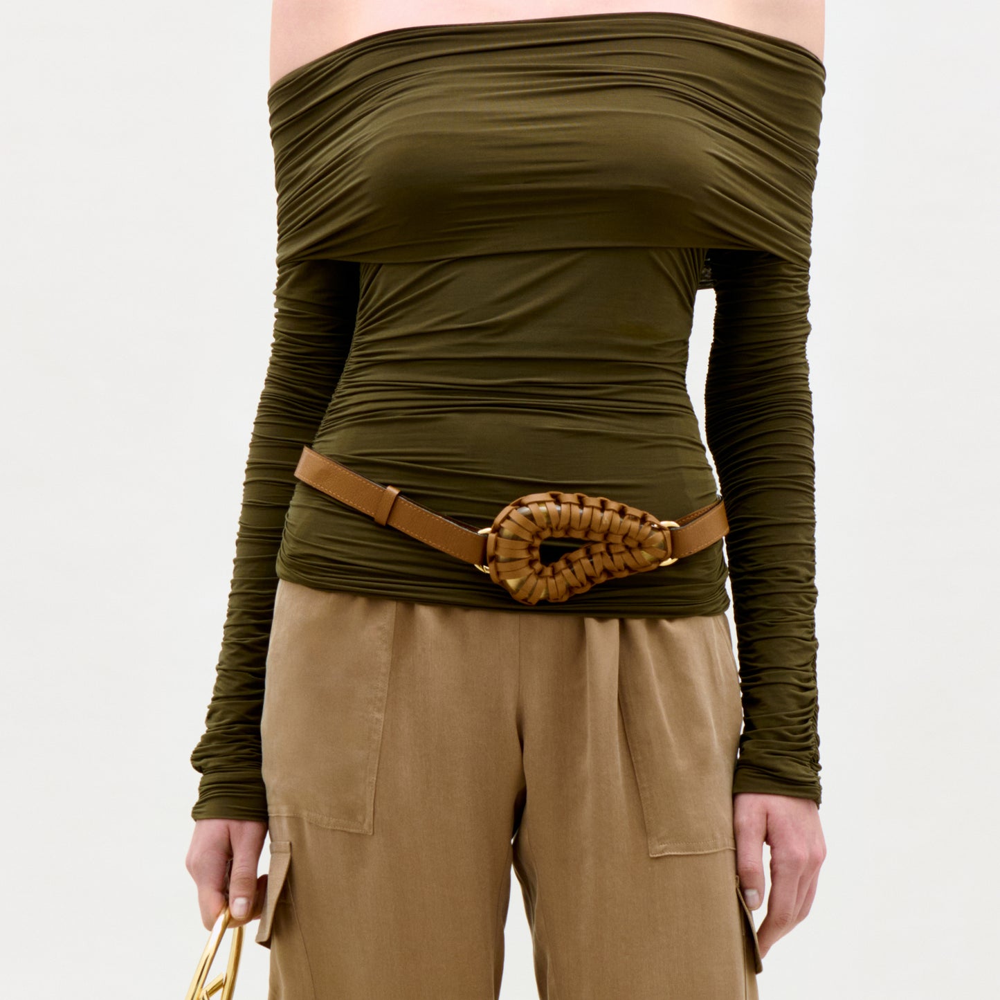 Person wearing an olive green off-shoulder top with a brown belt and beige pants on a white background