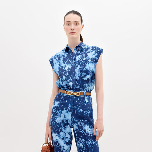 A woman wearing a blue tie-dye dress with a collar and sleeveless design, carrying a large navy blue bag on her shoulder, with her hair tied back in a ponytail, is showcasing the Rina Blouse Indigo exclusive pre-order item available until November 15th, 2024.