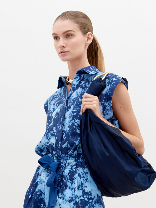 A woman wearing a blue tie-dye dress with a collar and sleeveless design, carrying a large navy blue bag on her shoulder, with her hair tied back in a ponytail, is showcasing the Rina Blouse Indigo exclusive pre-order item available until November 15th, 2024.