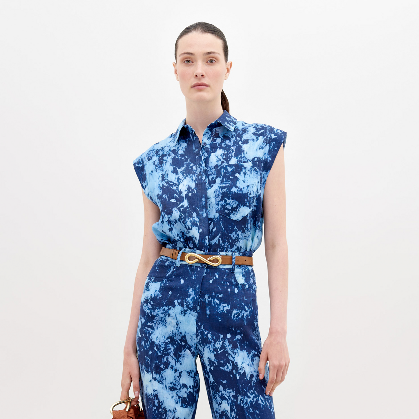 A woman wearing a blue tie-dye dress with a collar and sleeveless design, carrying a large navy blue bag on her shoulder, with her hair tied back in a ponytail, is showcasing the Rina Blouse Indigo exclusive pre-order item available until November 15th, 2024.