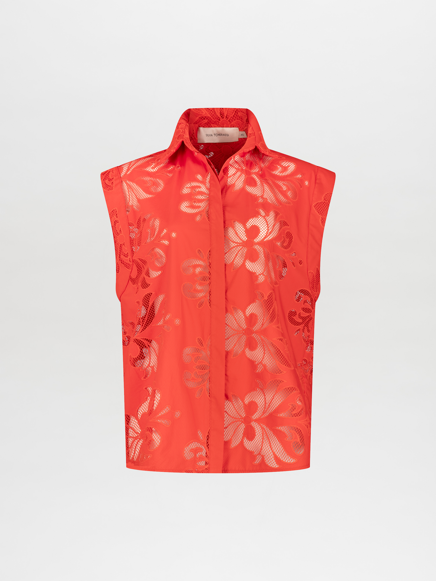 A person stands wearing a vibrant orange sleeveless top and matching pants with floral patterns, accessorized with a brown belt and holding a brown handbag. Pre-order the Rina Blouse Rouge Embroidery now to ensure it ships by the date of November 15th, 2024.