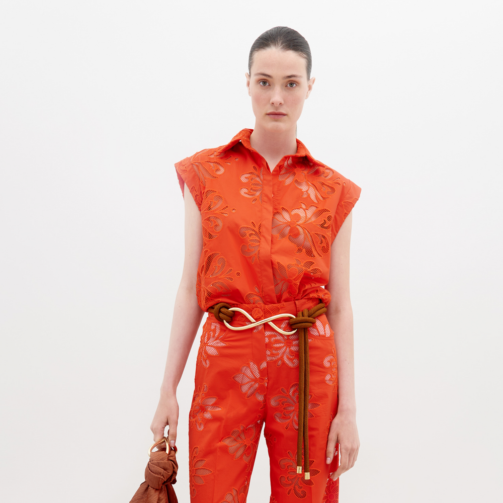 A person stands wearing a vibrant orange sleeveless top and matching pants with floral patterns, accessorized with a brown belt and holding a brown handbag. Pre-order the Rina Blouse Rouge Embroidery now to ensure it ships by the date of November 15th, 2024.
