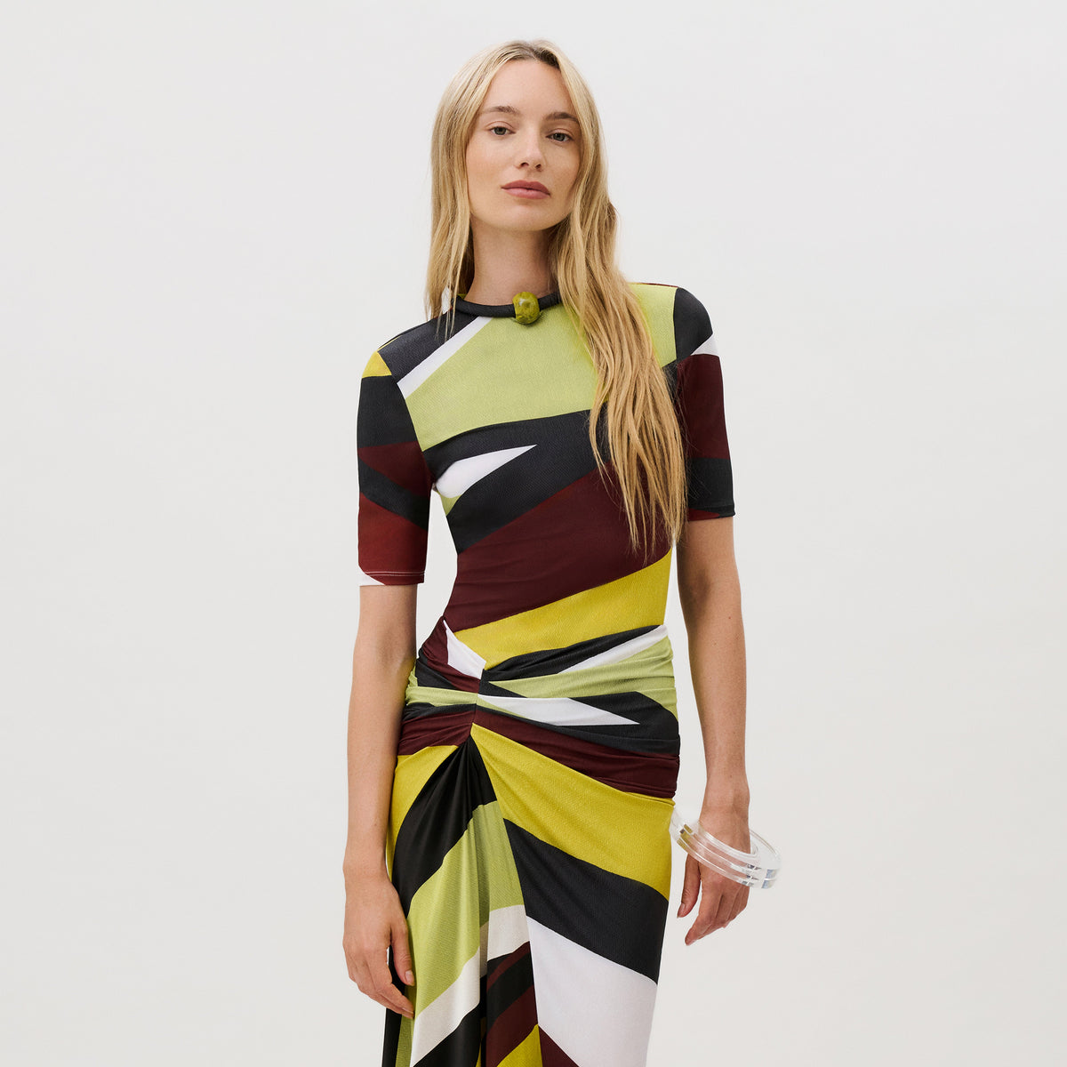A woman wearing a Rio Top featuring a slim fit, short-sleeved design with a geometric, multicolored abstract print, a rounded neckline, and a decorative stone detail at the collar.