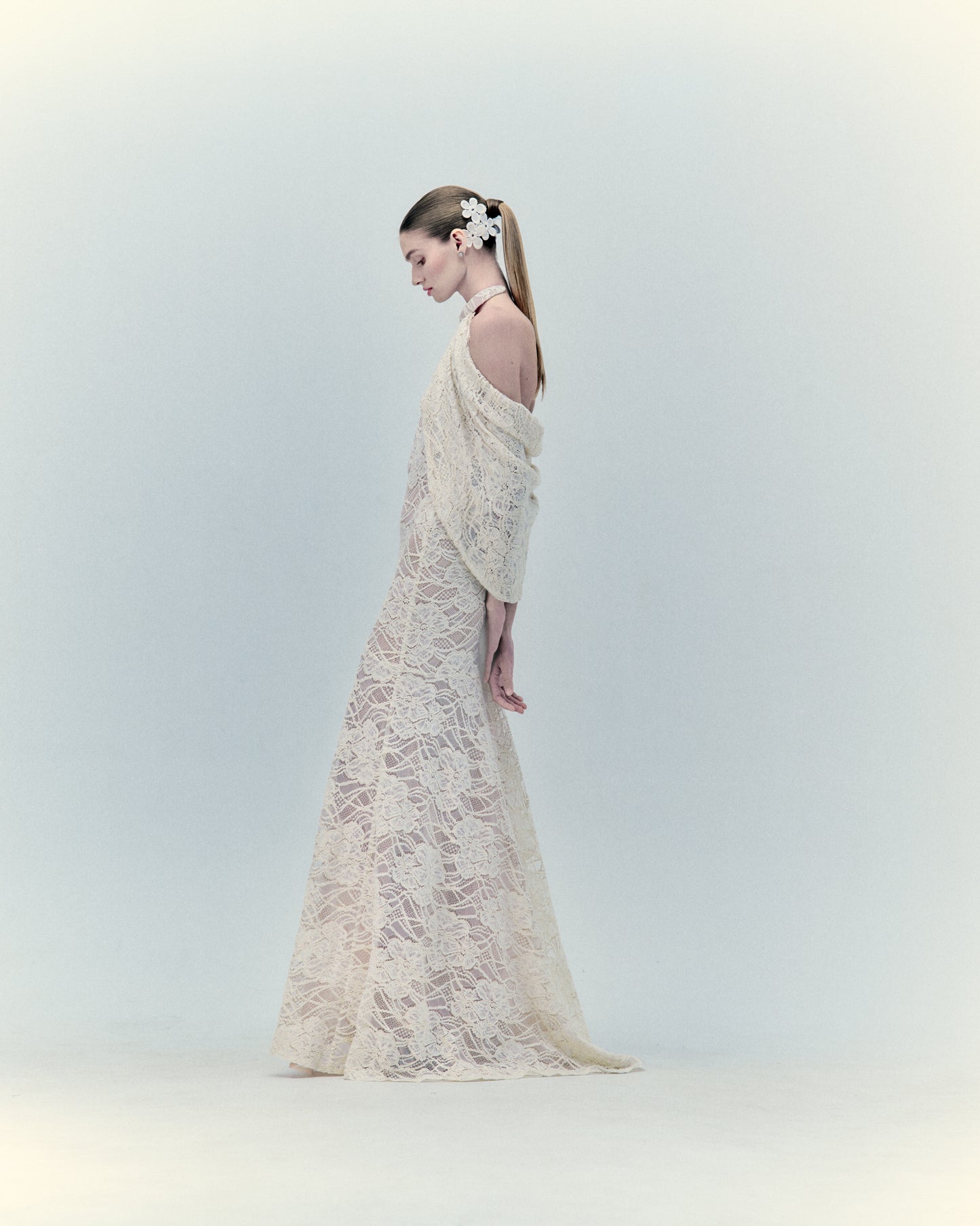 A woman in profile wearing the Rita Dress Beige, long and elegant with lace details, complemented by delicate floral accessories in her hair.