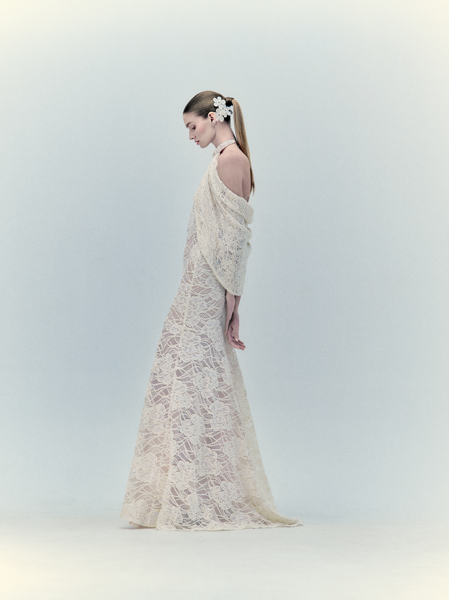 A woman in profile wearing the Rita Dress Beige, long and elegant with lace details, complemented by delicate floral accessories in her hair.