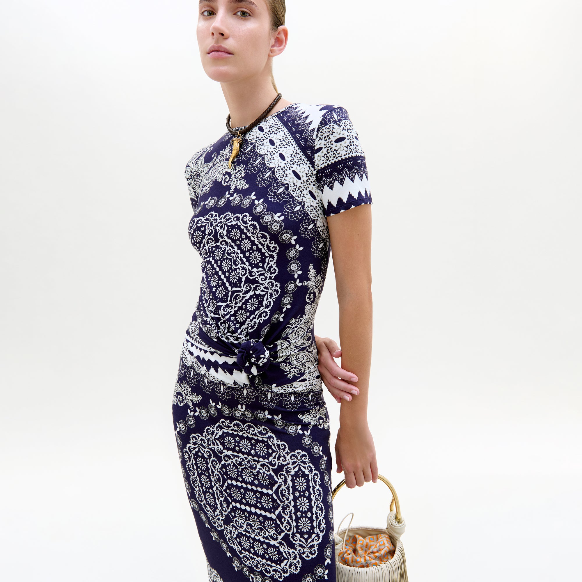 A person wearing the Riviera Dress, a fitted short-sleeve midi day dress in navy blue and white Mediterranean lace print, stands against a plain background, holding a small handbag with a round wooden handle and orange fabric lining.