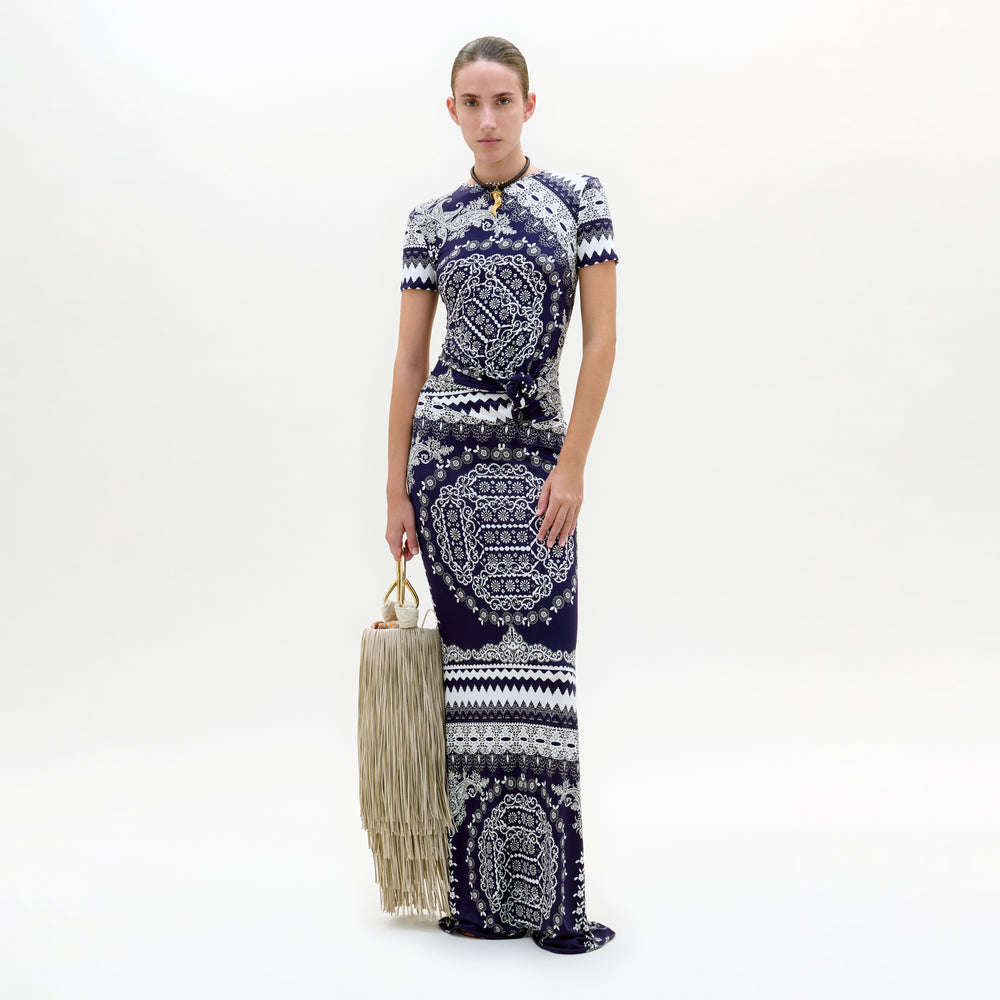 A woman stands against a white background wearing the Riviera Dress, a fitted, patterned navy and white maxi in Italian viscose. She holds a large beige fringed handbag and looks at the camera with a neutral expression.