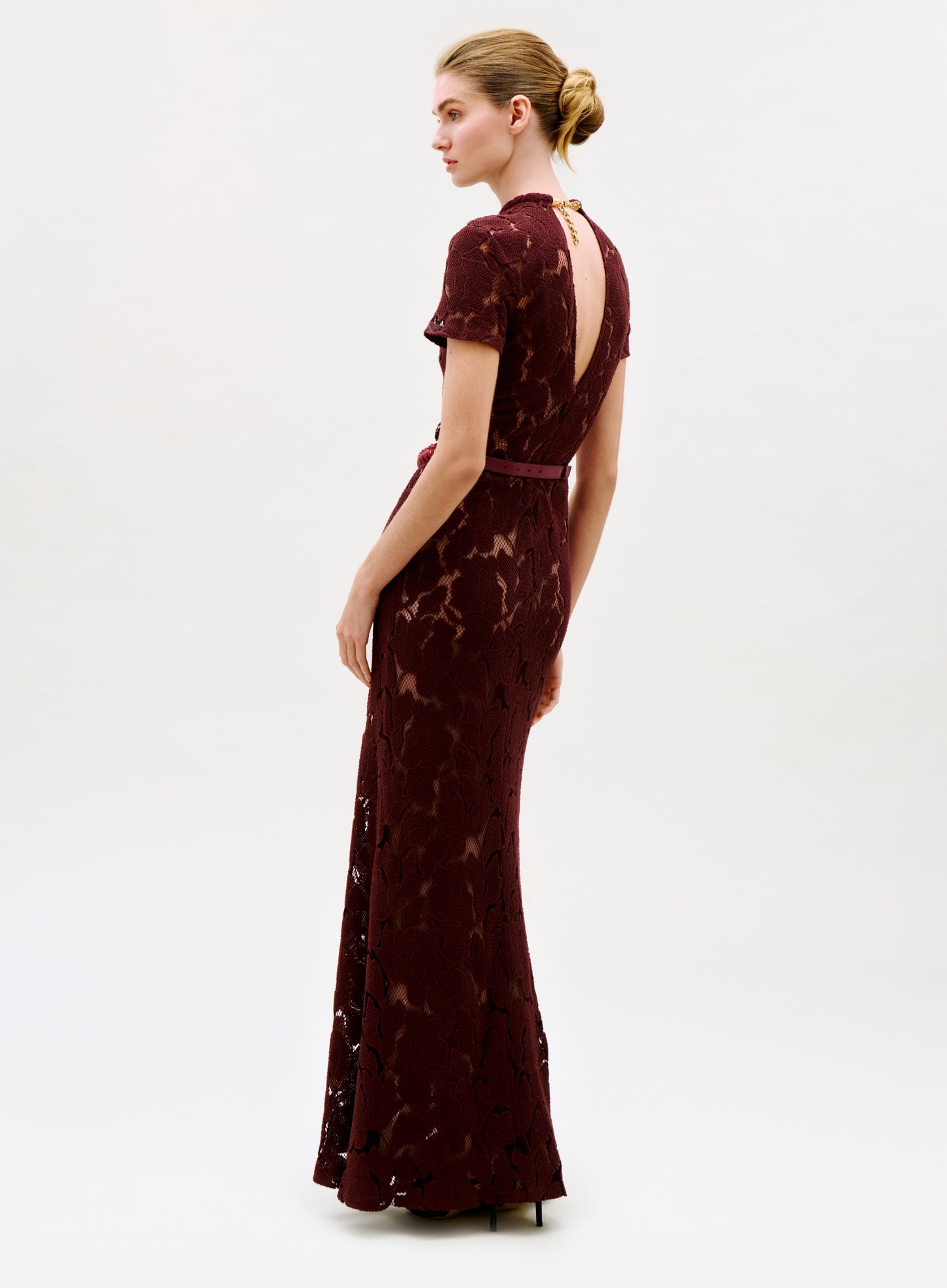 A woman with blonde hair in a low bun wears the Rochelle Dress—a floor-length, dark red lace gown with short sleeves and an open back—standing against a plain light background while looking over her shoulder.