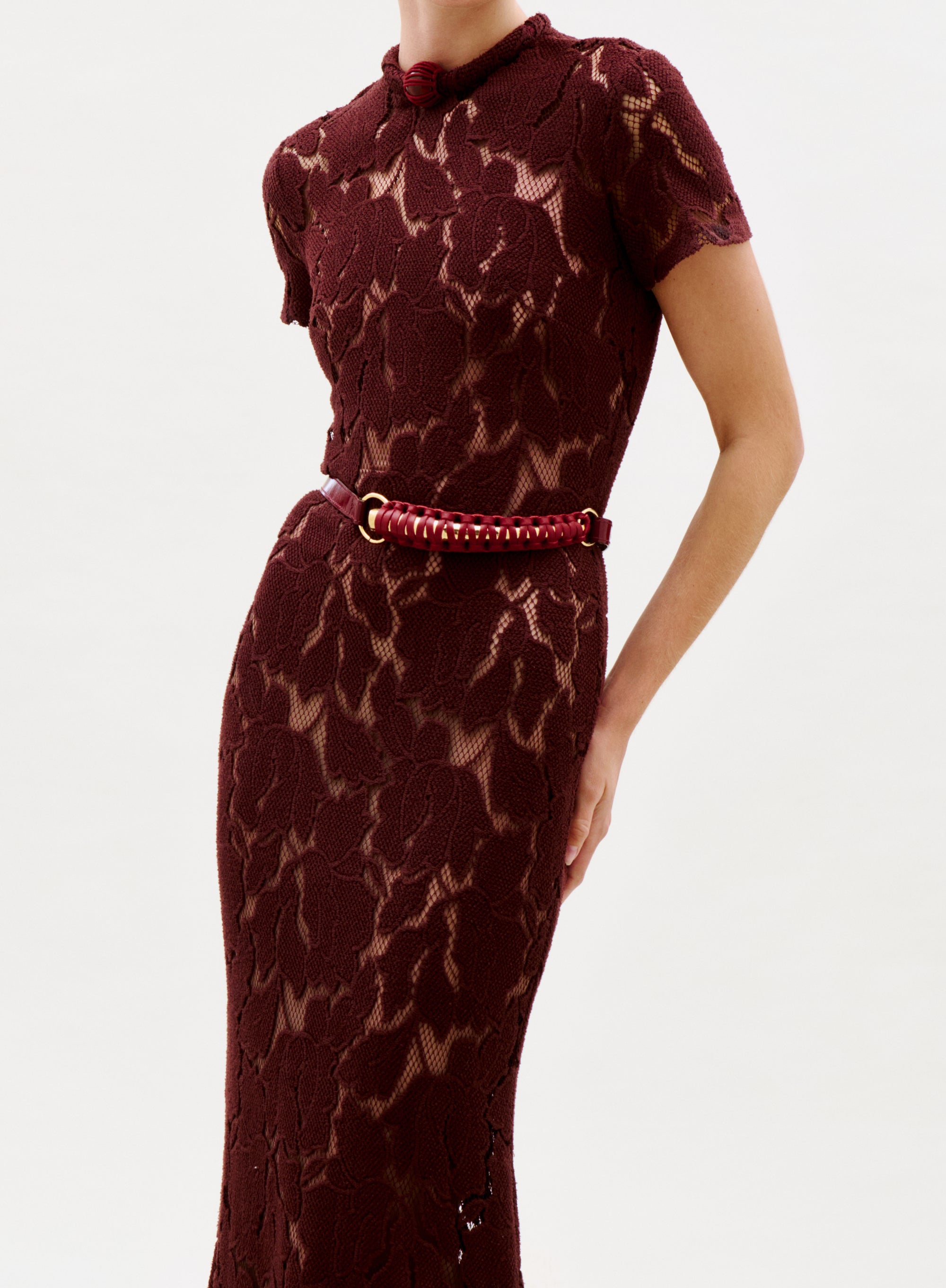 A person wears the Rochelle Dress—a fitted, short-sleeve burgundy lace dress with a matching belt, floral lace pattern, and high neckline—standing against a plain light background.