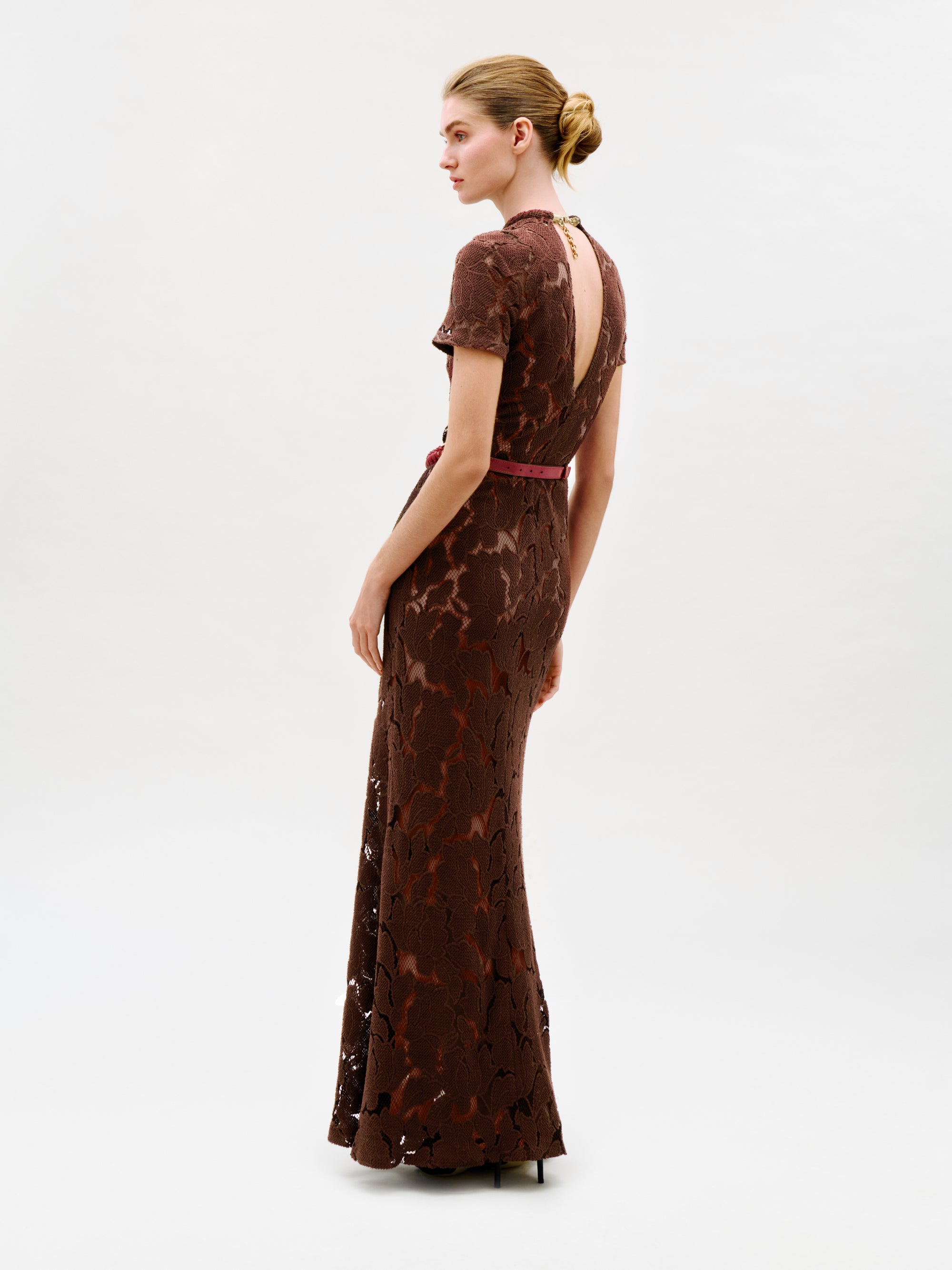 A woman stands sideways against a plain light background, looking to her left, wearing the Rochelle Dress—a long brown lace maxi dress with short sleeves and an open back. Crafted from luxurious Italian fabric and made in Colombia, she wears her hair in a neat bun and pairs the look with high heels.