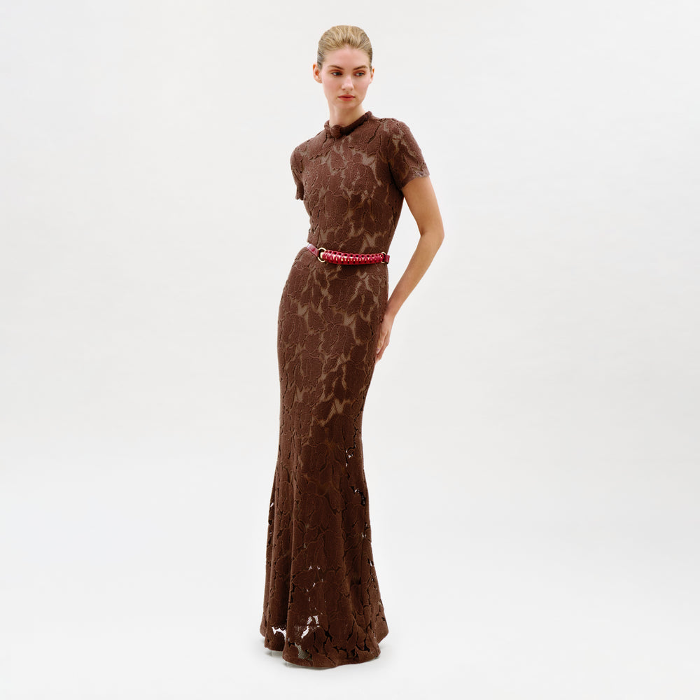 A woman stands against a plain white background wearing the Rochelle Dress, a fitted, floor-length brown lace maxi with short sleeves and a red belt, crafted from luxurious Italian fabric and made in Colombia.