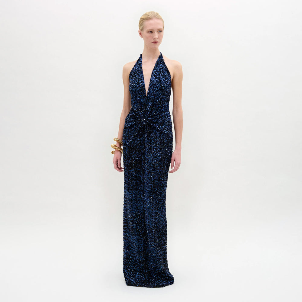 A person stands against a white background wearing the sleeveless, deep V-neck Rowan Dress in navy blue sequins with a gathered waist. Their blonde hair is pulled back and they accessorize with gold bracelets on their left wrist.