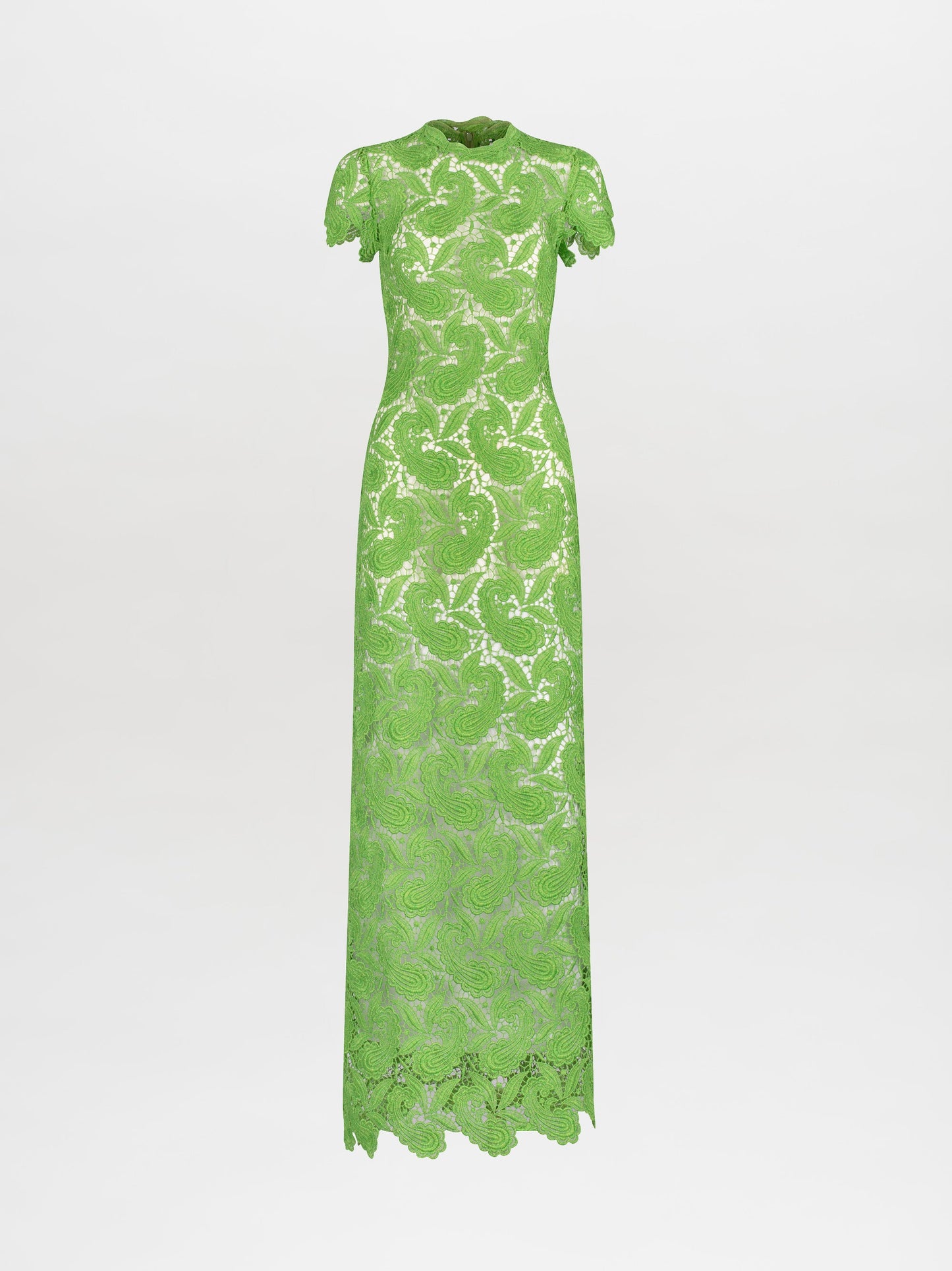 A person stands against a plain white background, wearing the Roxana Dress Green Guipure—a long, fitted lace dress with short sleeves. Hands are clasped behind the back. This elegant dress is available for pre-order now, with a scheduled ship date of February 15th, 2025.