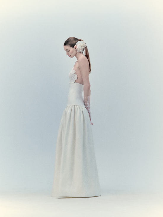 A woman in a Ruth Dress Pearl gown, elegant white with floral embellishments, wearing a decorative hairpiece, stands side profile against a light backdrop.