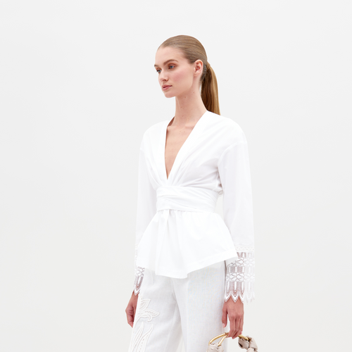 Person wearing the Saanvi Blouse White with lace detail and coordinating white pants, holding a small handbag, looking off to the side against a plain background. Don't miss your chance to pre-order this stunning outfit by the ship-by date of February 15th, 2025.