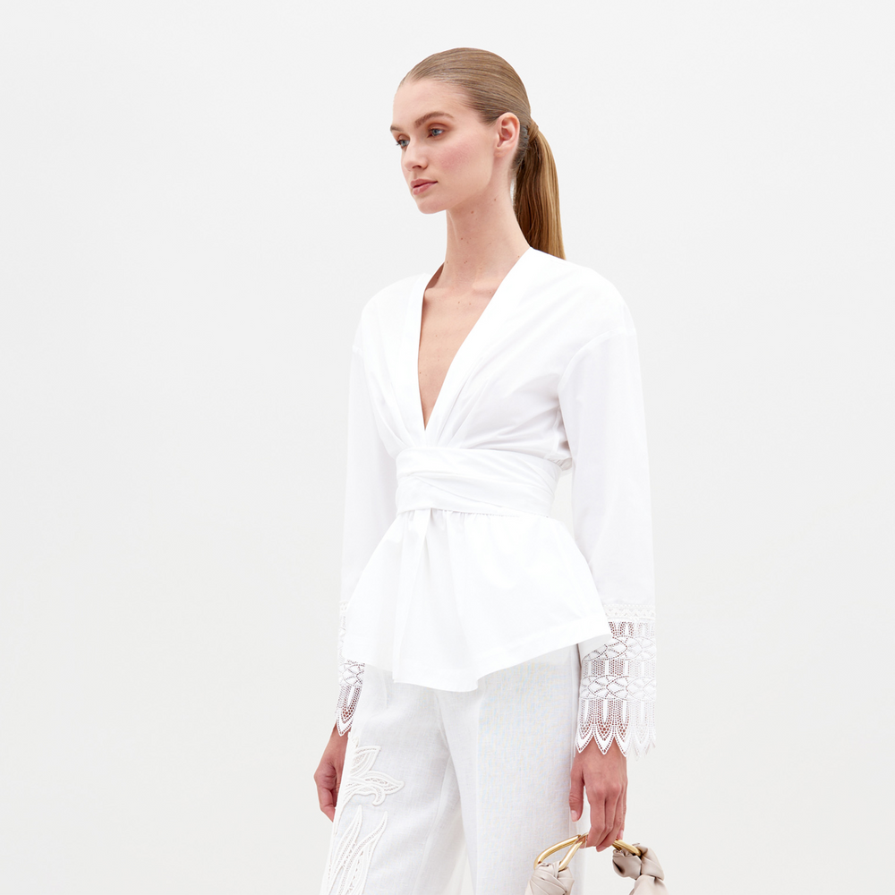 Person wearing the Saanvi Blouse White with lace detail and coordinating white pants, holding a small handbag, looking off to the side against a plain background. Don't miss your chance to pre-order this stunning outfit by the ship-by date of February 15th, 2025.