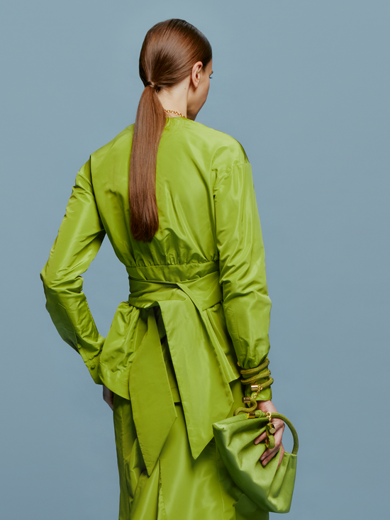 Wearing the elegant Saanvi Blouse in Lime, paired with a gold necklace and matching handbag, a woman stands against a plain blue background, exuding effortless style as she embraces the calm of September 2nd.