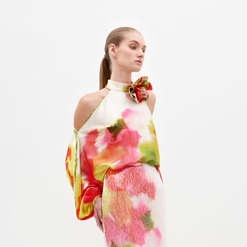 A person dressed in a vibrant floral Safira Blouse with intricate appliqué detailing stands against a plain backdrop.