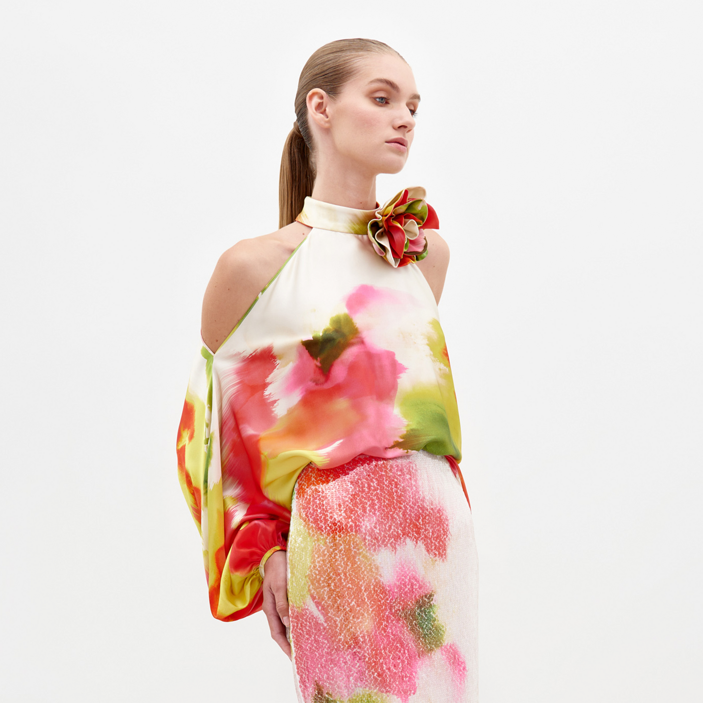 A woman models the Safira Blouse, featuring a colorful watercolor floral pattern, halter neckline, and a stylish floral appliqué on the shoulder, set against a plain white background.
