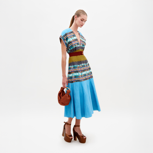 A woman stands wearing a Sakura Dress in Aqua Olive Plaid, featuring a V-neck design with multicolored patterns. She holds a brown handbag and wears brown platform heels. Available for pre-order, this look will ship by February 15th, 2025.