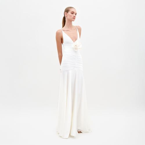 A woman in the Sana Dress White, featuring a sleeveless design with a deep V-neck and floral detail at the waist, stands against a plain white background. Pre-order now to ensure delivery by February 15th, 2025.