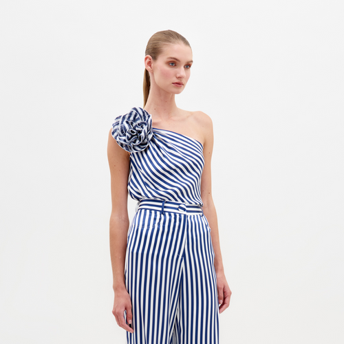 A woman stands wearing the Sebastiana Blouse in white and blue stripes, featuring a one-shoulder design with a large flower detail on the shoulder, against a plain white background. This chic outfit is available for pre-order now and will ship by February 15th, 2025.