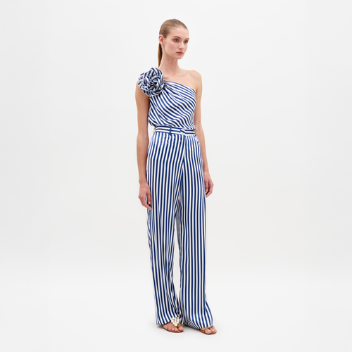 A woman stands wearing the Emine Pant White Blue Stripes, a one-shoulder jumpsuit featuring blue and white stripes and a large shoulder ruffle, available for pre-order until February 15th, 2025.