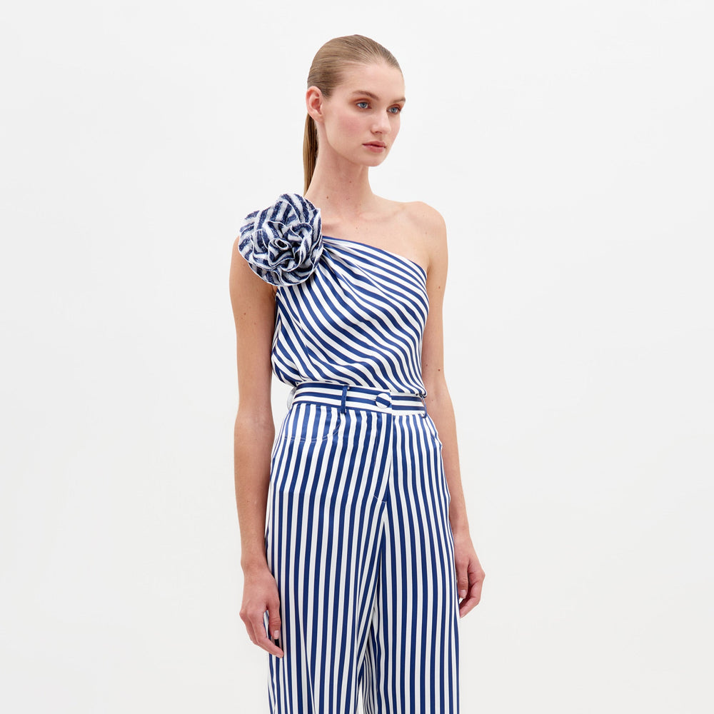 A woman stands wearing the Sebastiana Blouse in white and blue stripes, featuring a one-shoulder design with a large flower detail on the shoulder, against a plain white background. This chic outfit is available for pre-order now and will ship by February 15th, 2025.