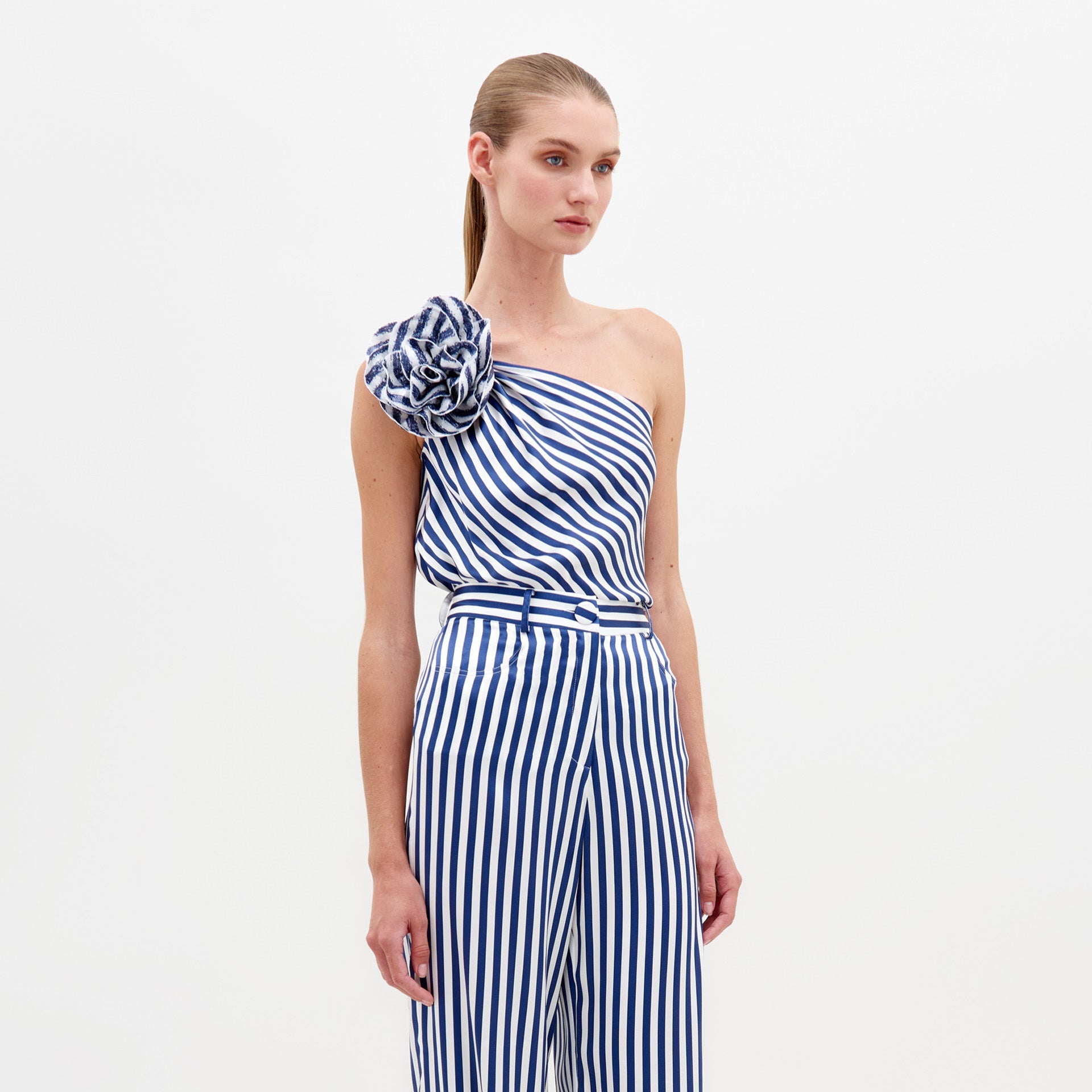 A woman stands wearing the Sebastiana Blouse in white and blue stripes, featuring a one-shoulder design with a large flower detail on the shoulder, against a plain white background. This chic outfit is available for pre-order now and will ship by February 15th, 2025.