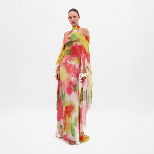 A person is standing in the Serenela Dress Spring Blooms, a colorful, flowing dress with a halter neck and floral patterns, holding a small white flower bouquet. The dress is ready to be pre-ordered for the February 15th ship-by date.