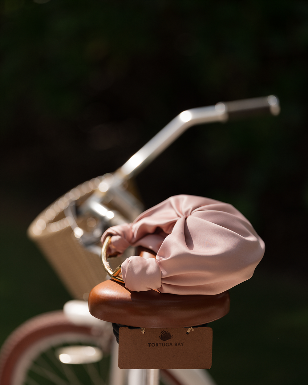 Brown bicycle seat with a pink satin Sofia Bag resting on it, against a blurred background. A 