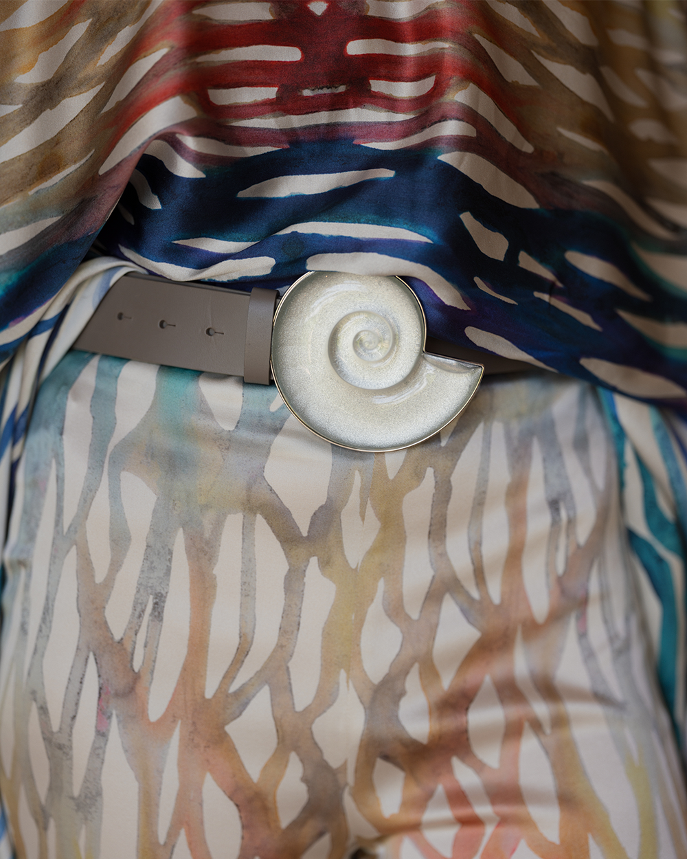 Abstract pattern fabric in earth tones, blended with deep blue and red, drapes from the waist. A large, pearl-white spiral belt buckle serves as a focal point.