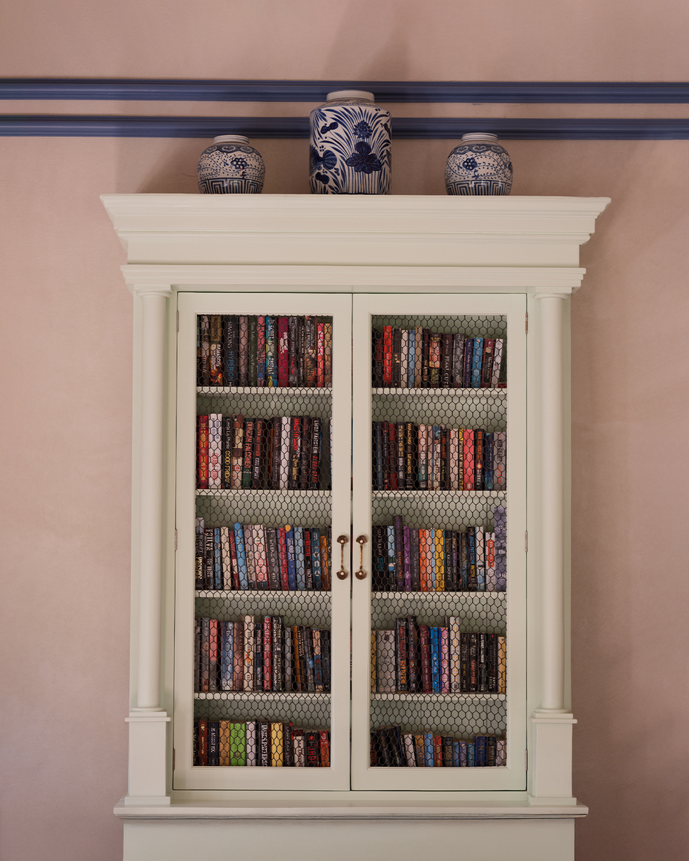 A white cabinet filled with colorful, patterned books behind mesh doors is topped with three blue and white decorative vases, set against a peach wall.