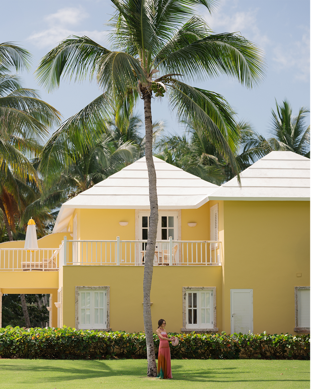 A woman in a colorful dress stands near a palm tree in front of a bright yellow house with a white roof. The scene is sunny and tropical.