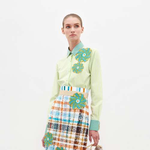 A woman stands against a plain background wearing the Shao Blouse Green Stripes, adorned with colorful floral embellishments, and a multicolored plaid skirt. She holds a small item in her left hand, now available for pre-order and expected to ship by February 15th, 2025.