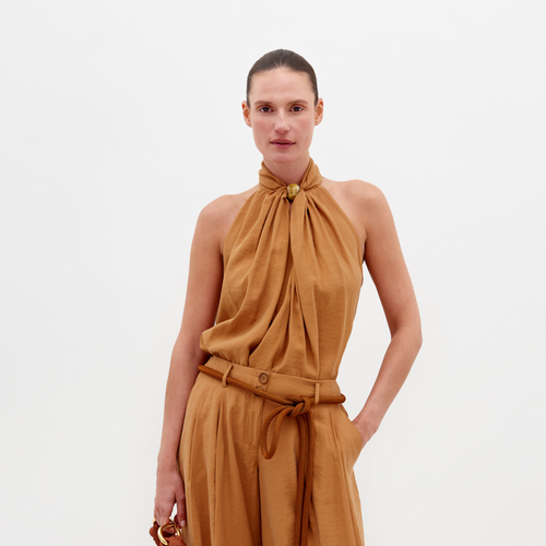 A person wearing the Shela Blouse Camel sleeveless high-neck top with matching wide-leg trousers, accessorized with a belt and holding a small object, stands against a plain white background. Pre-order now to ensure it ships by February 15th, 2025.