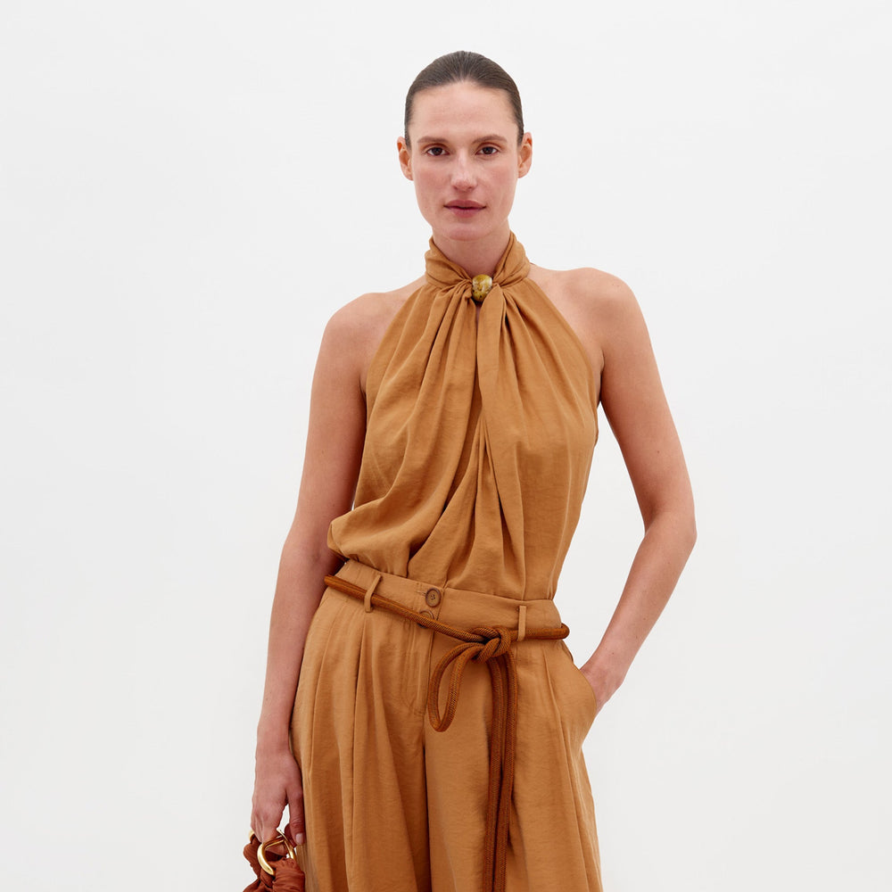 A person wearing the Shela Blouse Camel sleeveless high-neck top with matching wide-leg trousers, accessorized with a belt and holding a small object, stands against a plain white background. Pre-order now to ensure it ships by February 15th, 2025.