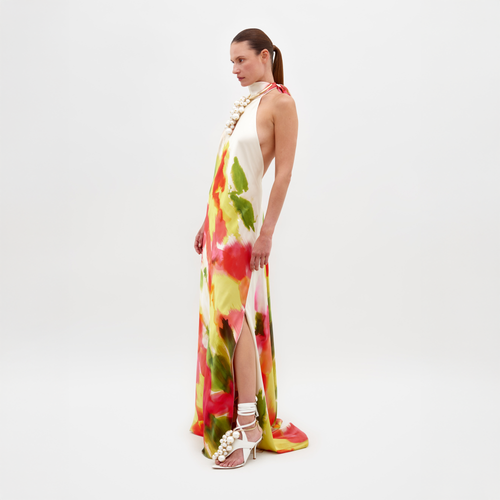 A person in the Sherry Dress Spring Blooms, featuring a multicolored print and a thigh-high slit, poses against a plain background. They are wearing white high-heeled sandals and a pearl necklace. Remember, this stunning outfit is available for pre-order and will ship by February 15th.